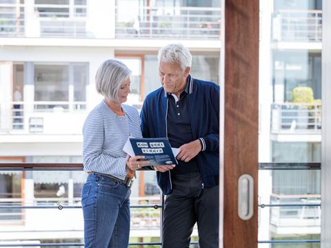 Parret står tæt sammen i en ny lejlighed og gennemgår en brochure med ekstra info. Den lyse baggrund skaber en rolig atmosfære.