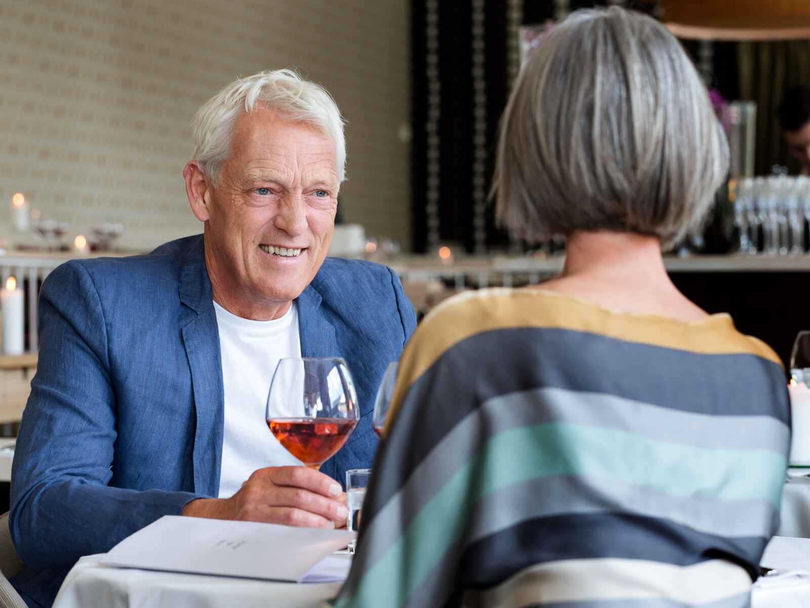 Manden smiler varmt med et glas vin i hånden, mens parret nyder en afslappet og intim middag i en elegant restaurant.