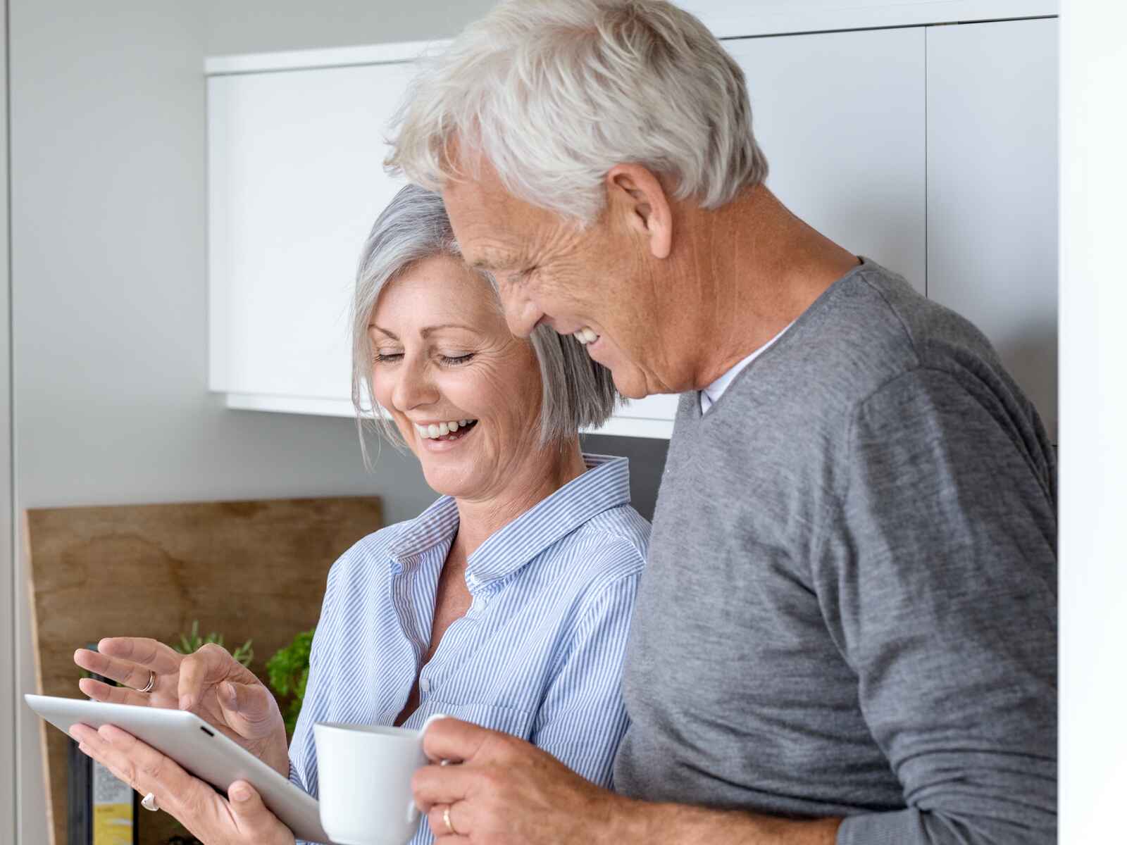 Parret smiler sammen, mens de kigger på en tablet i køkkenet. Kvinden holder en kop, og omgivelserne er lyse og moderne med en hyggelig atmosfære.