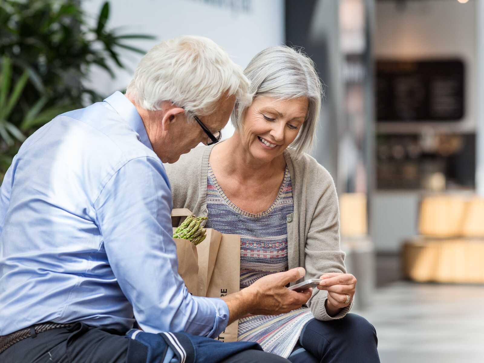 Ældre par sidder udendørs med indkøbsposer og ser på en telefon. Kvinden smiler, og manden er fokuseret. Grønne planter i baggrunden.