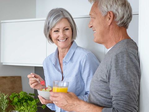 Ældre par sidder i køkkenet, kvinden spiser frugt, og manden holder et glas juice. Friske krydderurter og mad ses på køkkenbordet.