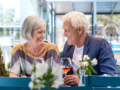 Ældre par sidder ved et restaurantbord, smiler til hinanden og nyder vin. Blomster på bordet og moderne omgivelser i baggrunden.