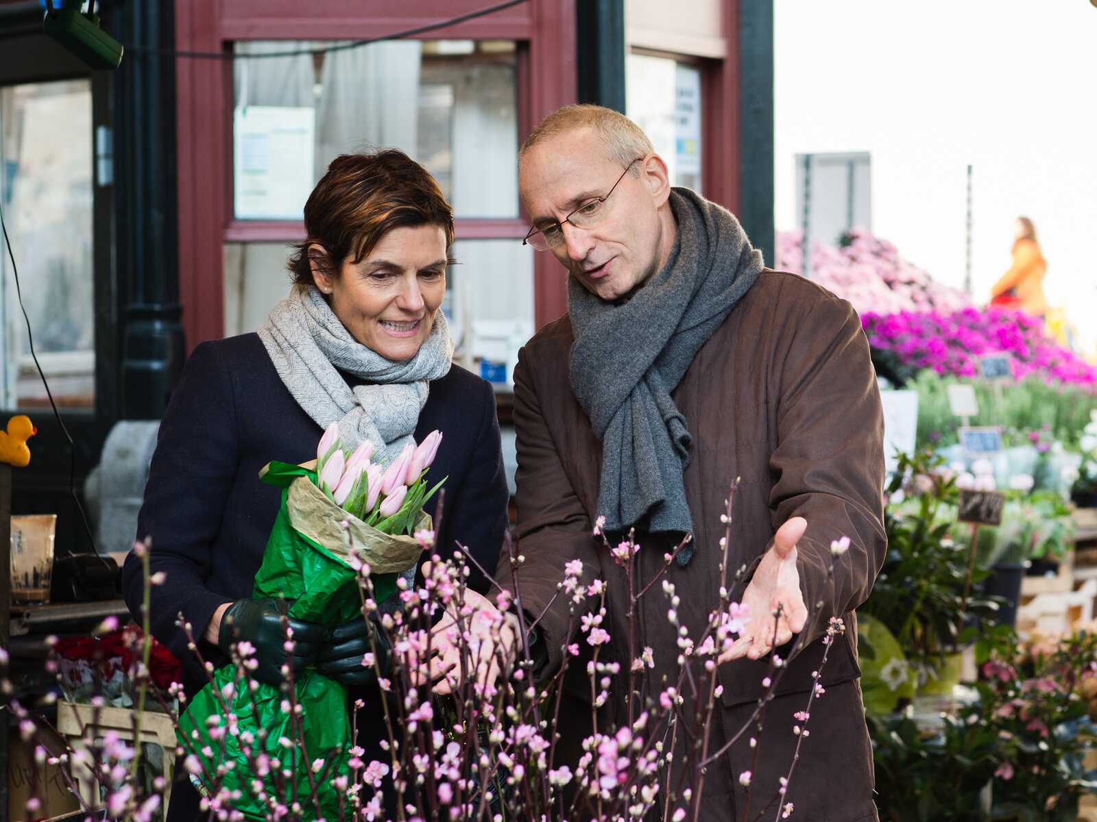 Par står ved en blomsterbod og kigger på blomstergrene. Kvinden holder en buket tulipaner, og manden peger på pink blomster.