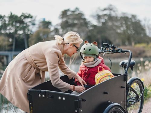 Mor hjælper sin datter med at spænde sikkerhedsselen i en ladcykel. Barnet bærer cykelhjelm og rygsæk, omgivelserne er udendørs med natur.