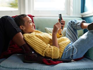 Kvinde i gul striktrøje smiler, mens hun ligger på sofaen og bruger en telefon med oplader i et hyggeligt opholdsrum.