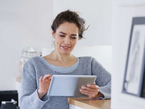 Kvinde i grå sweater står i køkkenet og kigger på en tablet. Baggrunden er lys med køkkenredskaber og glasbeholdere.
