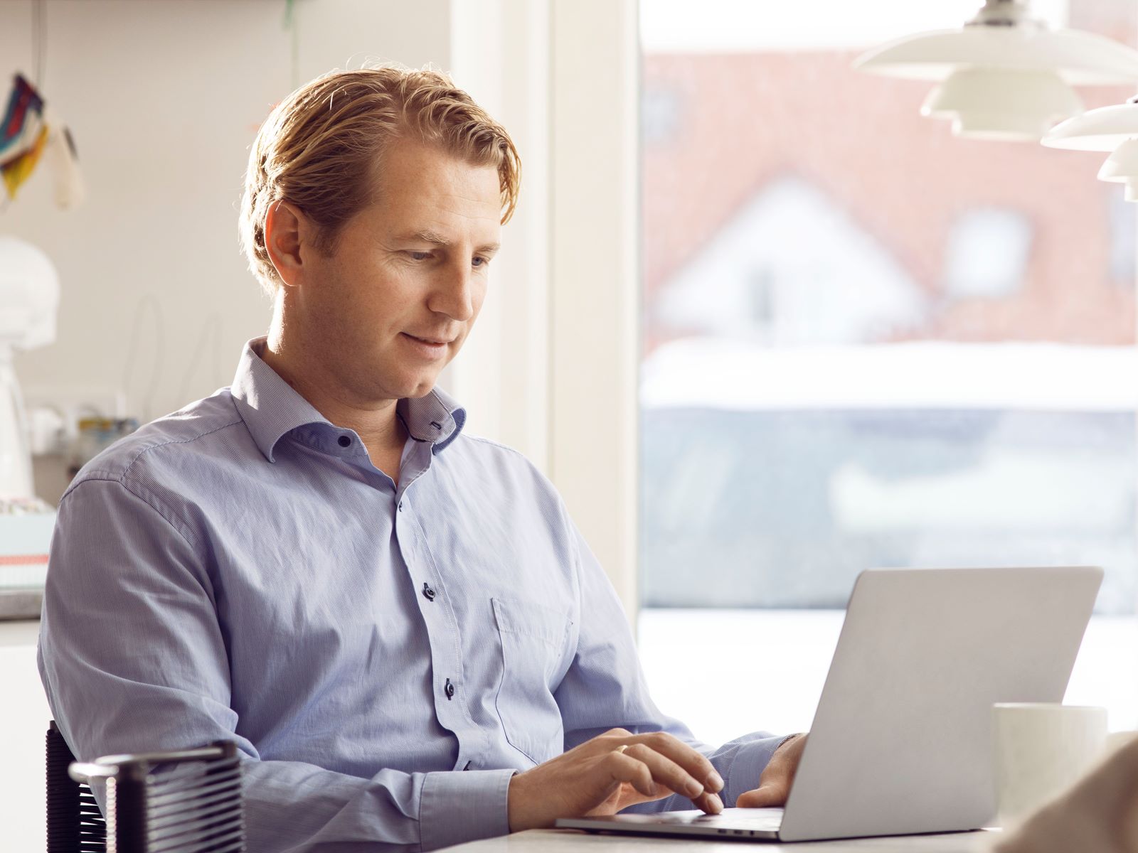 Mand i blå skjorte arbejder fokuseret på sin bærbare computer ved et vindue med naturligt lysindfald i baggrunden.