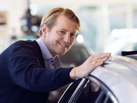 Smilende mand i forretningsdragt læner sig kærligt mod en ny bil i et showroom.