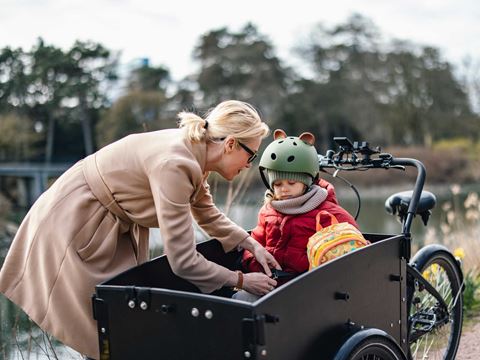 En mor spænder sin datter fast i en ladcykel, hvor datteren bærer en grøn hjelm med ører og en farverig rygsæk.