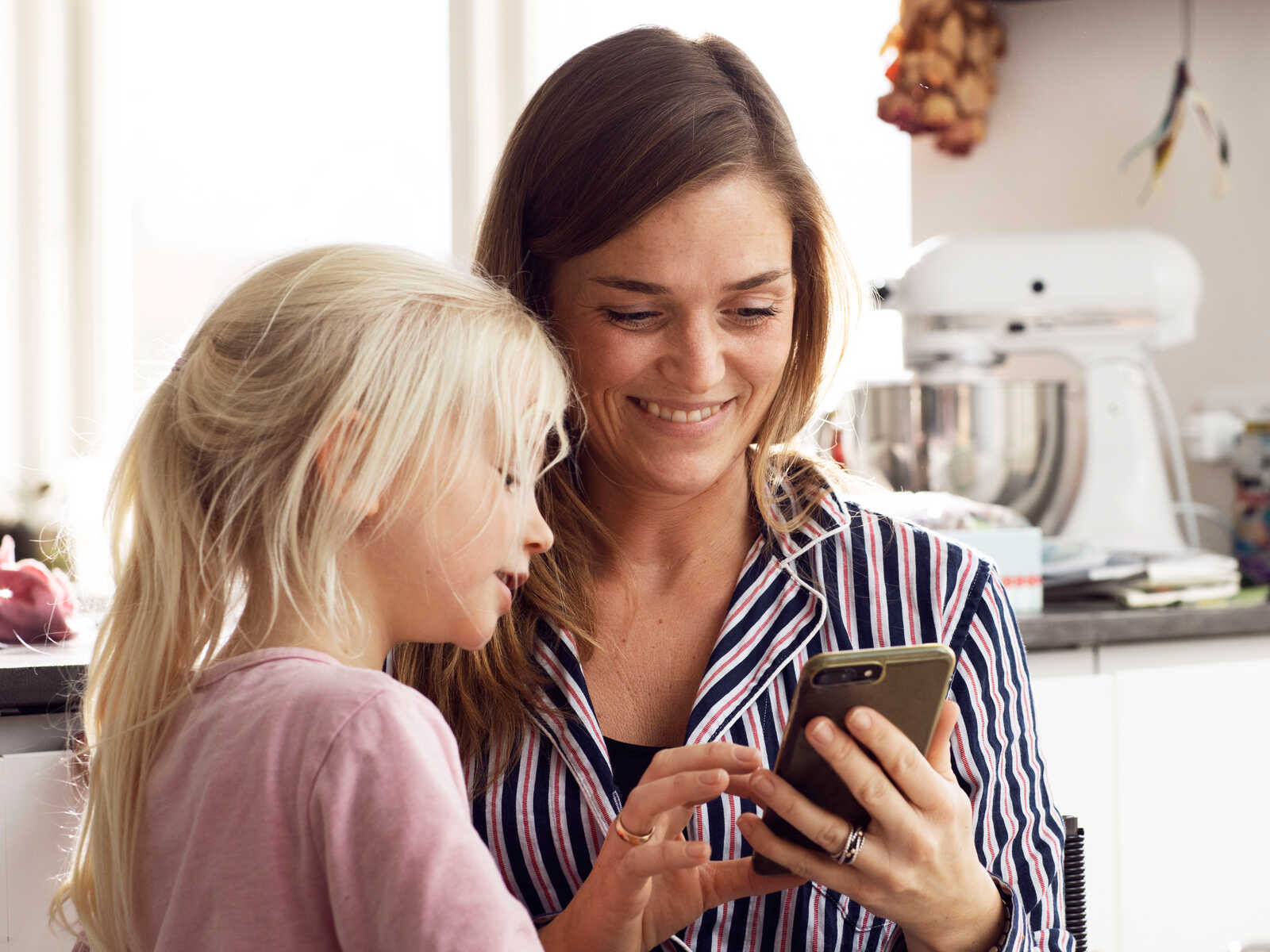 En mor og hendes datter sidder sammen i køkkenet og smiler, mens de kigger på noget på en smartphone.