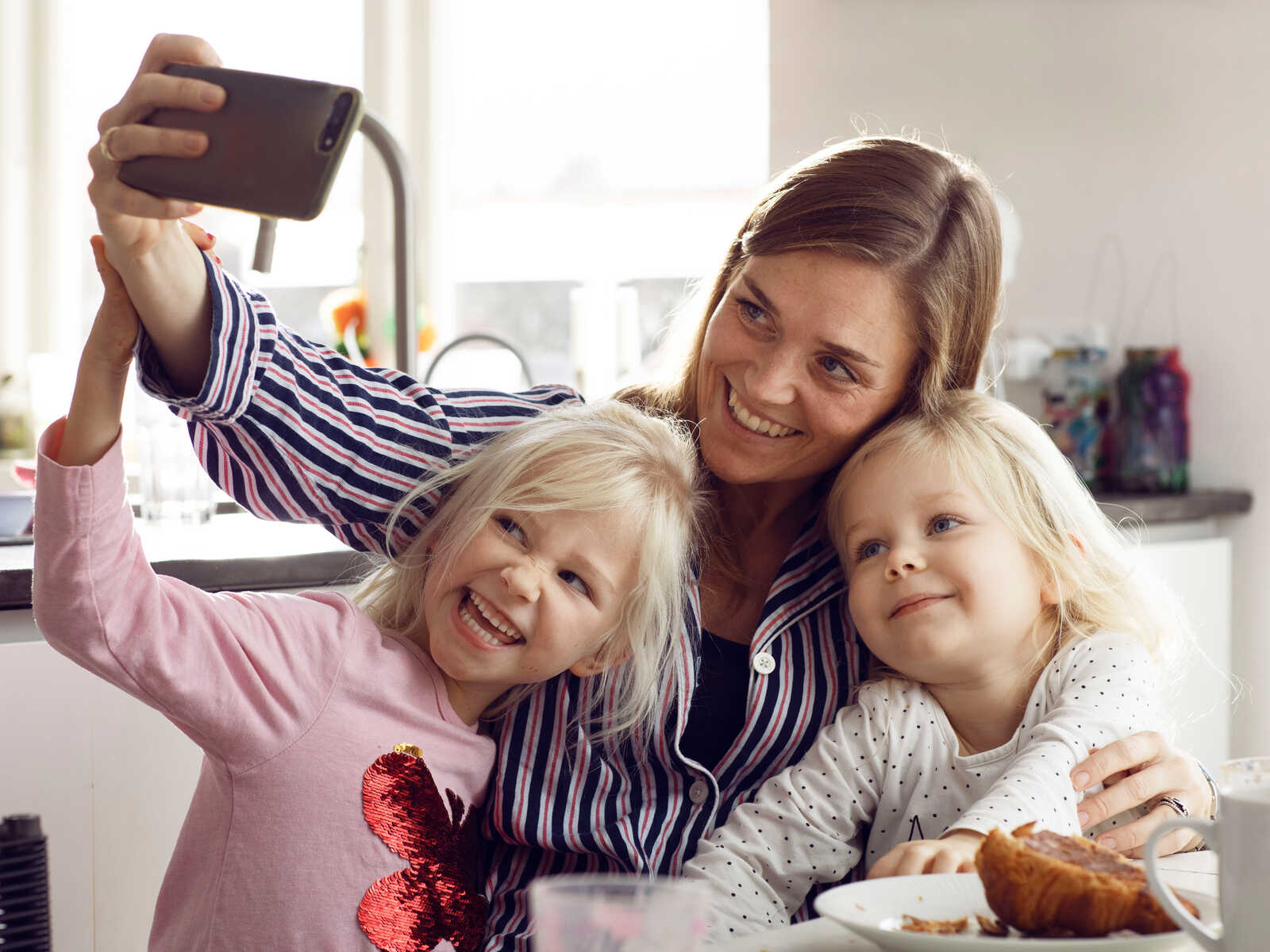 En mor tager en sjov selfie med sine to døtre i køkkenet, mens de alle smiler og laver ansigter.