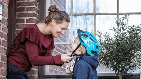 En mor fastgør sin søns cykelhjelm, mens de står udendørs foran et murstenshus og smiler til hinanden.