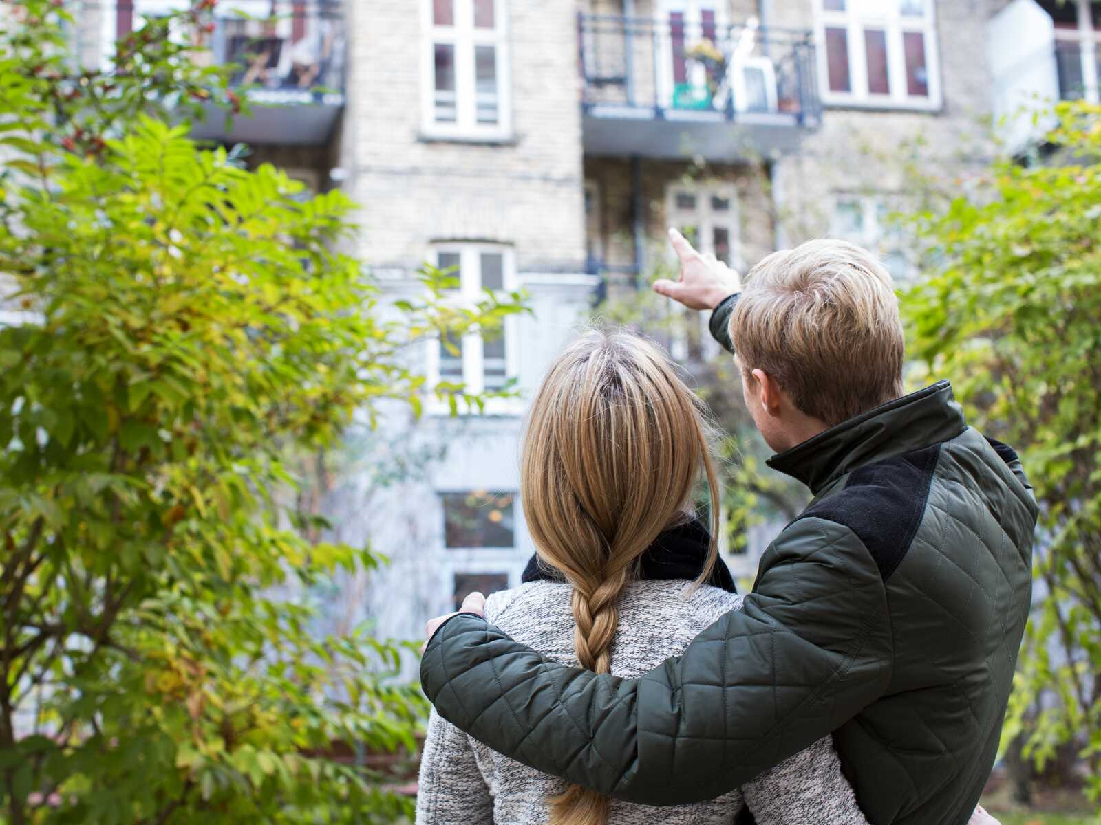 Et par står i en gårdhave og peger mod en lejlighedsbalkon, mens de kigger op på bygningen sammen.