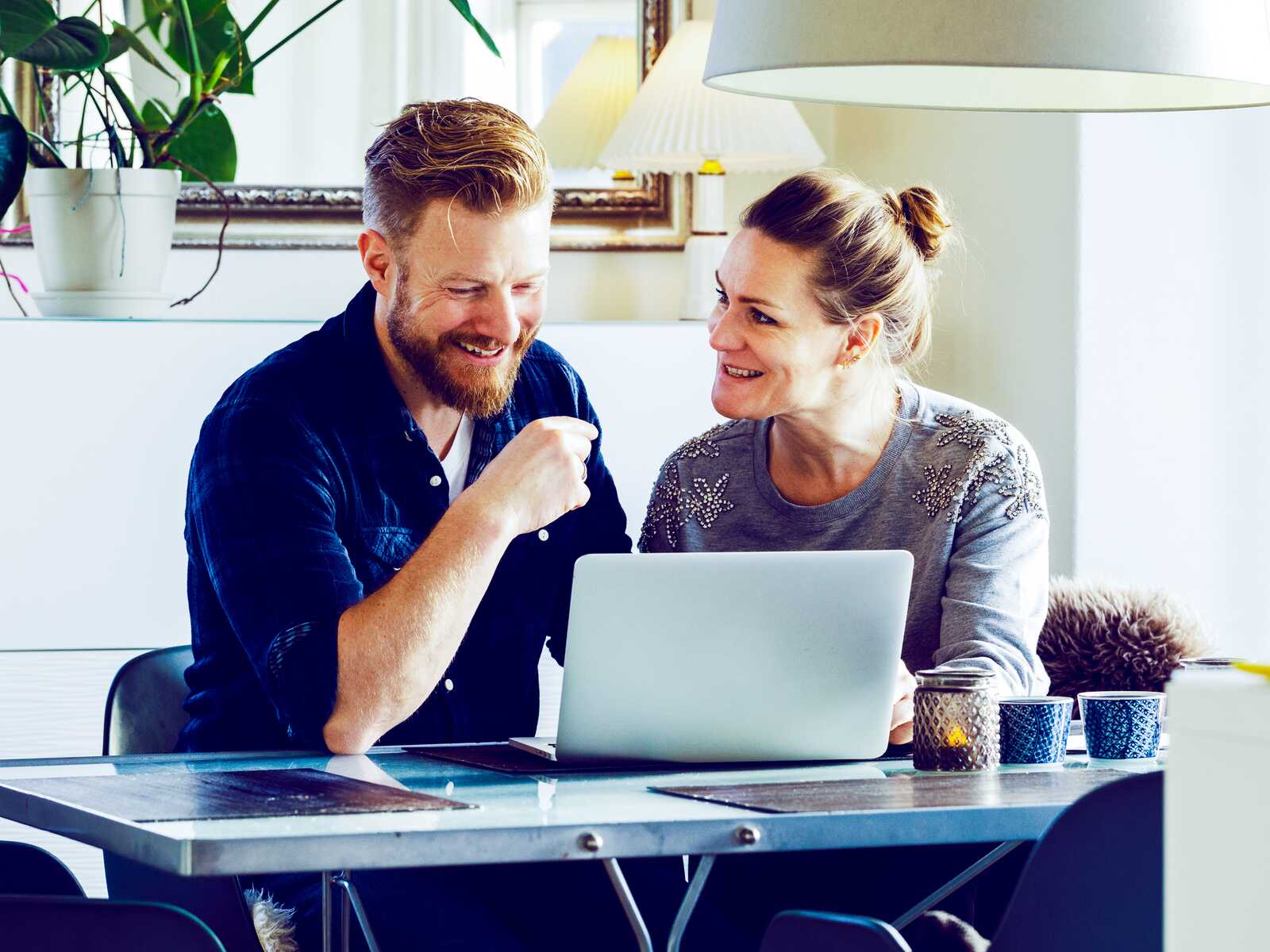 Et smilende par sidder ved et bord i stuen og arbejder sammen på en bærbar computer, mens de diskuterer noget.