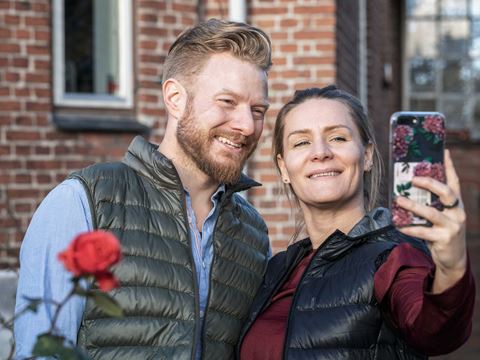 Et smilende par står udendørs foran et murstenshus og tager en selfie sammen.