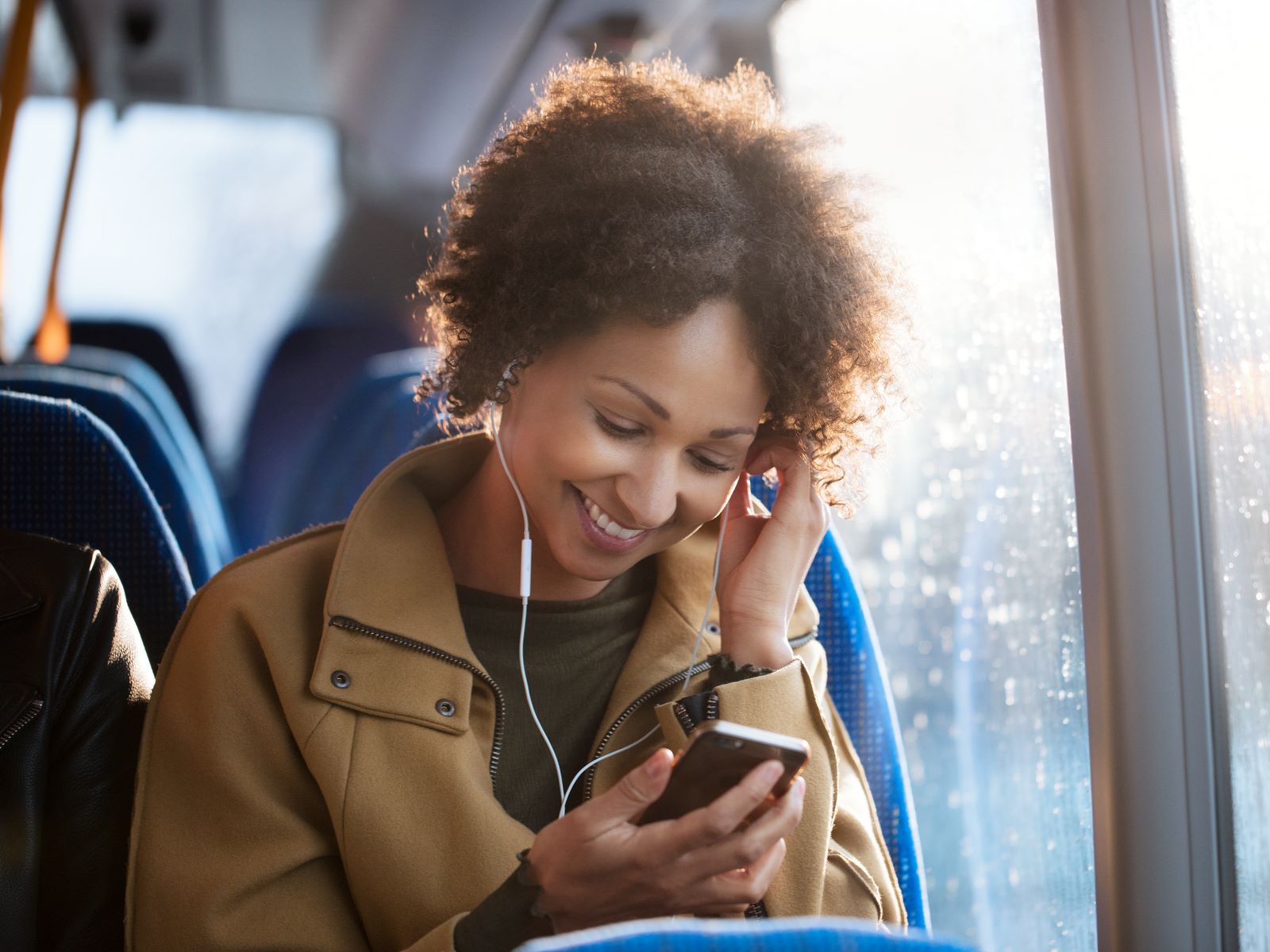 En kvinde sidder i en bus med høretelefoner på og smiler, mens hun kigger på sin telefon.