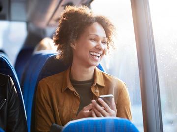 En kvinde sidder i en bus og smiler bredt, mens hun holder sin telefon og kigger ud af vinduet.