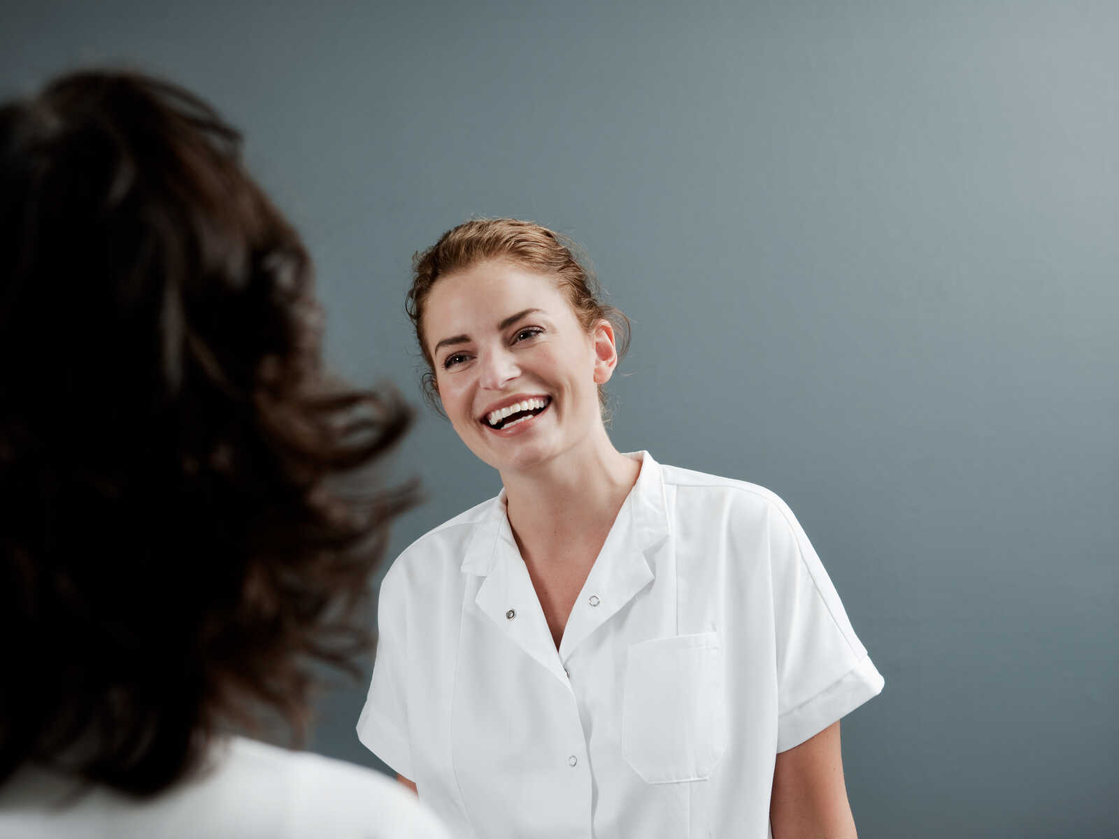 En smilende kvinde iført en hvid skjorte ser glad ud, mens hun taler med en person uden for billedet.
