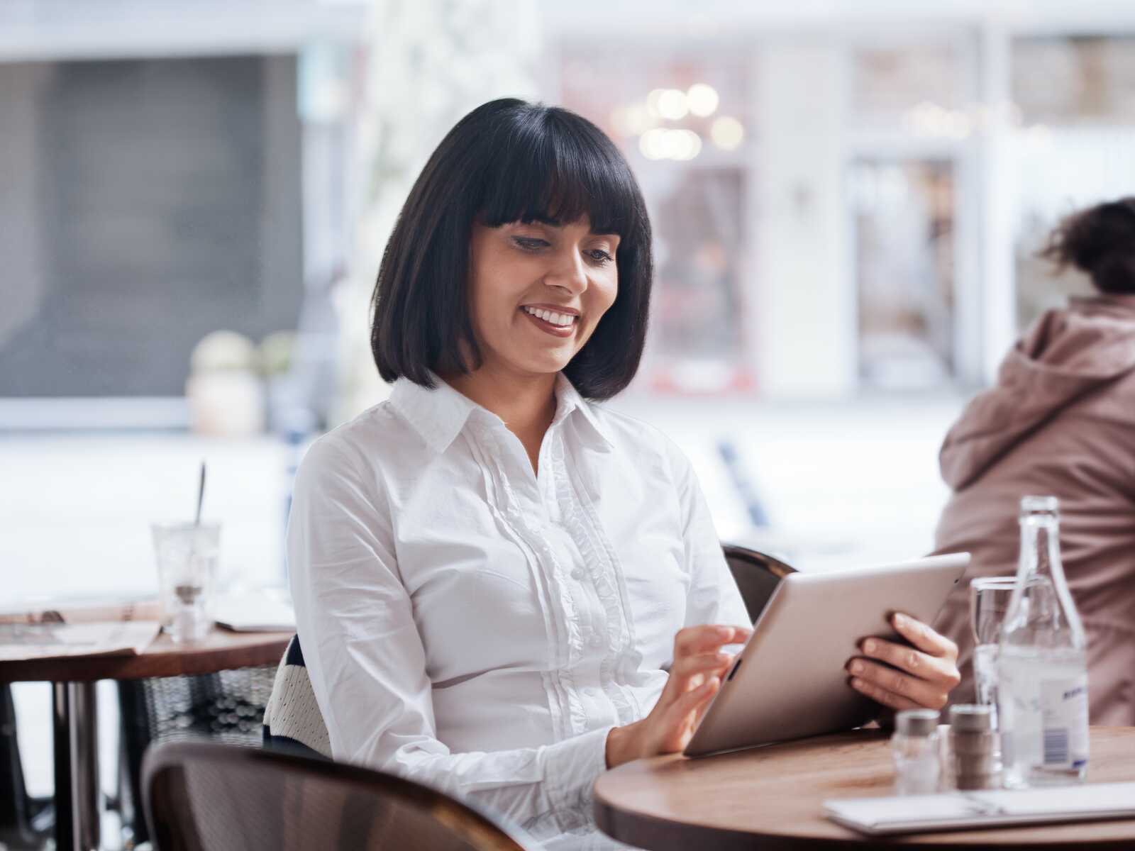 En kvinde sidder på en café og smiler, mens hun bruger en tablet ved et bord.
