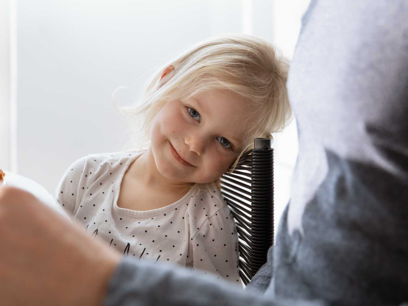 Et lille barn med lyst hår smiler sødt, mens hun hviler hovedet på en stol og kigger roligt frem.