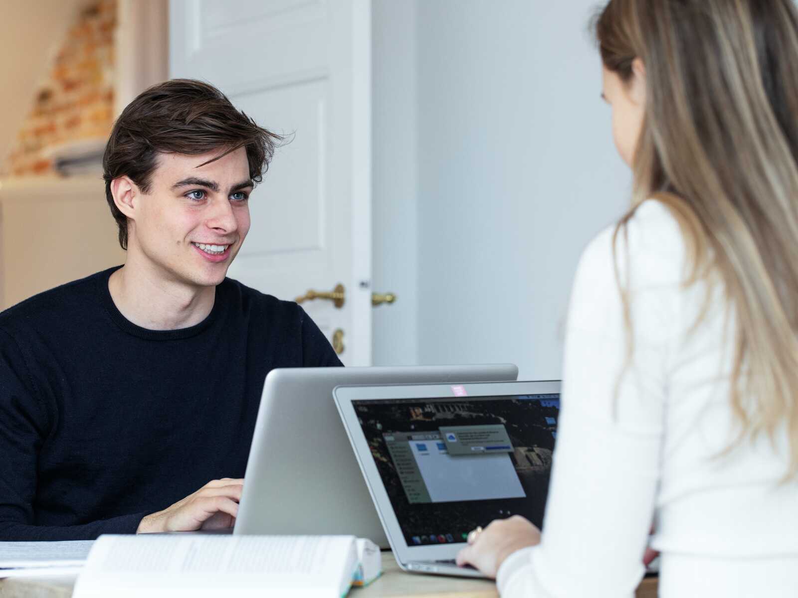 En ung mand smiler, mens han arbejder ved en bærbar computer sammen med en studiepartner i et lyst rum.