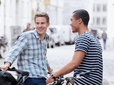 To unge mænd står og smiler, mens de snakker sammen på gaden. Den ene står ved sin cykel.