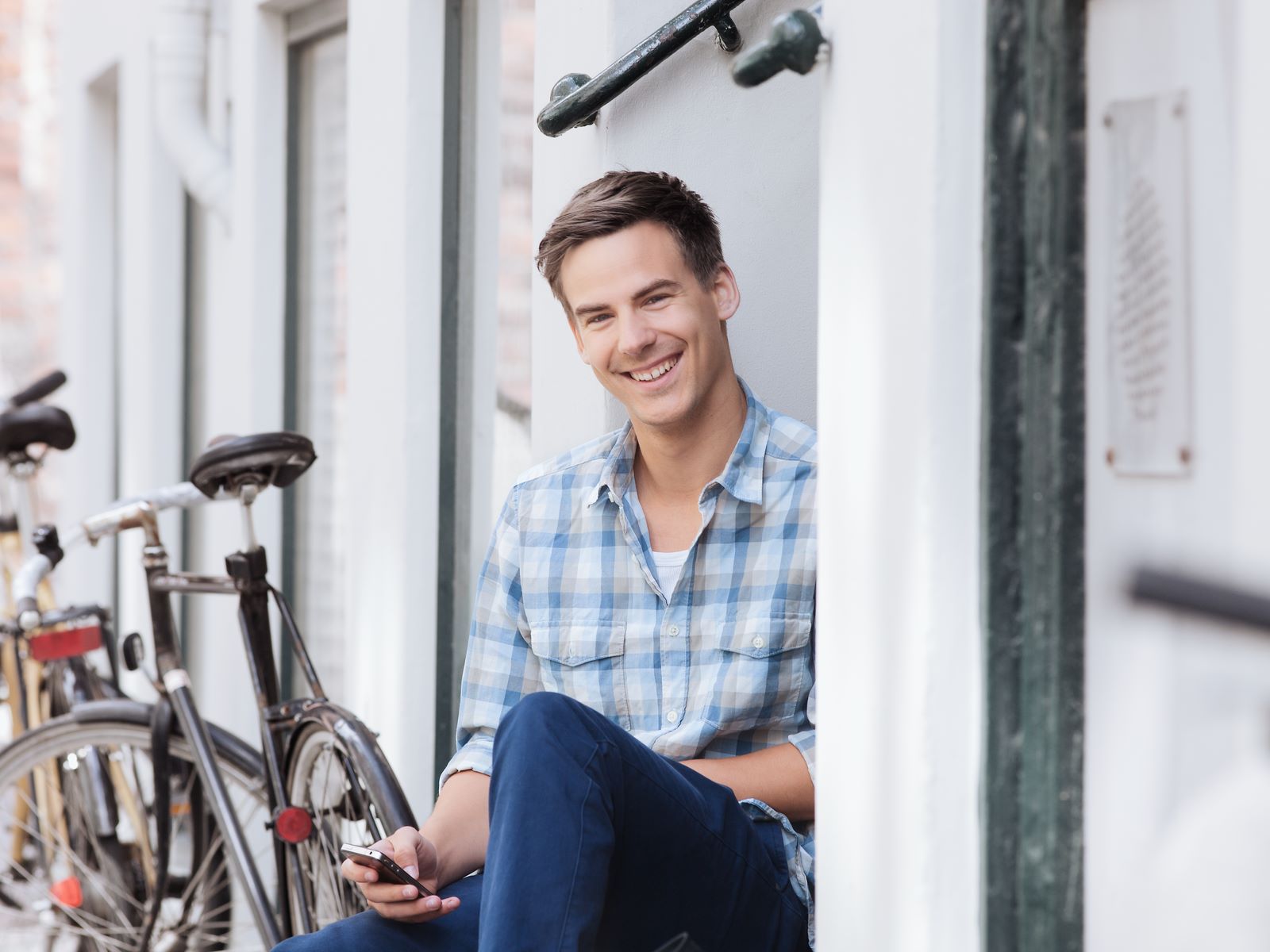 En ung mand i ternet skjorte sidder afslappet på en trappesten ved siden af cykler, smilende og kigger på sin telefon.