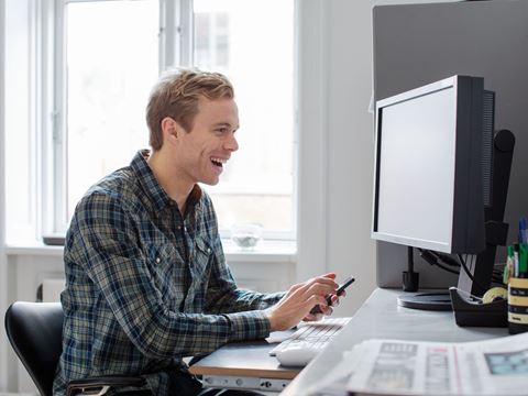 Begejstret ung mand smiler, mens han arbejder ved sin computer og holder sin telefon i hånden.