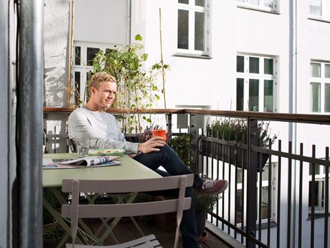 ung mand sidder på altan på en stol og smiler ned i sin telefon 
