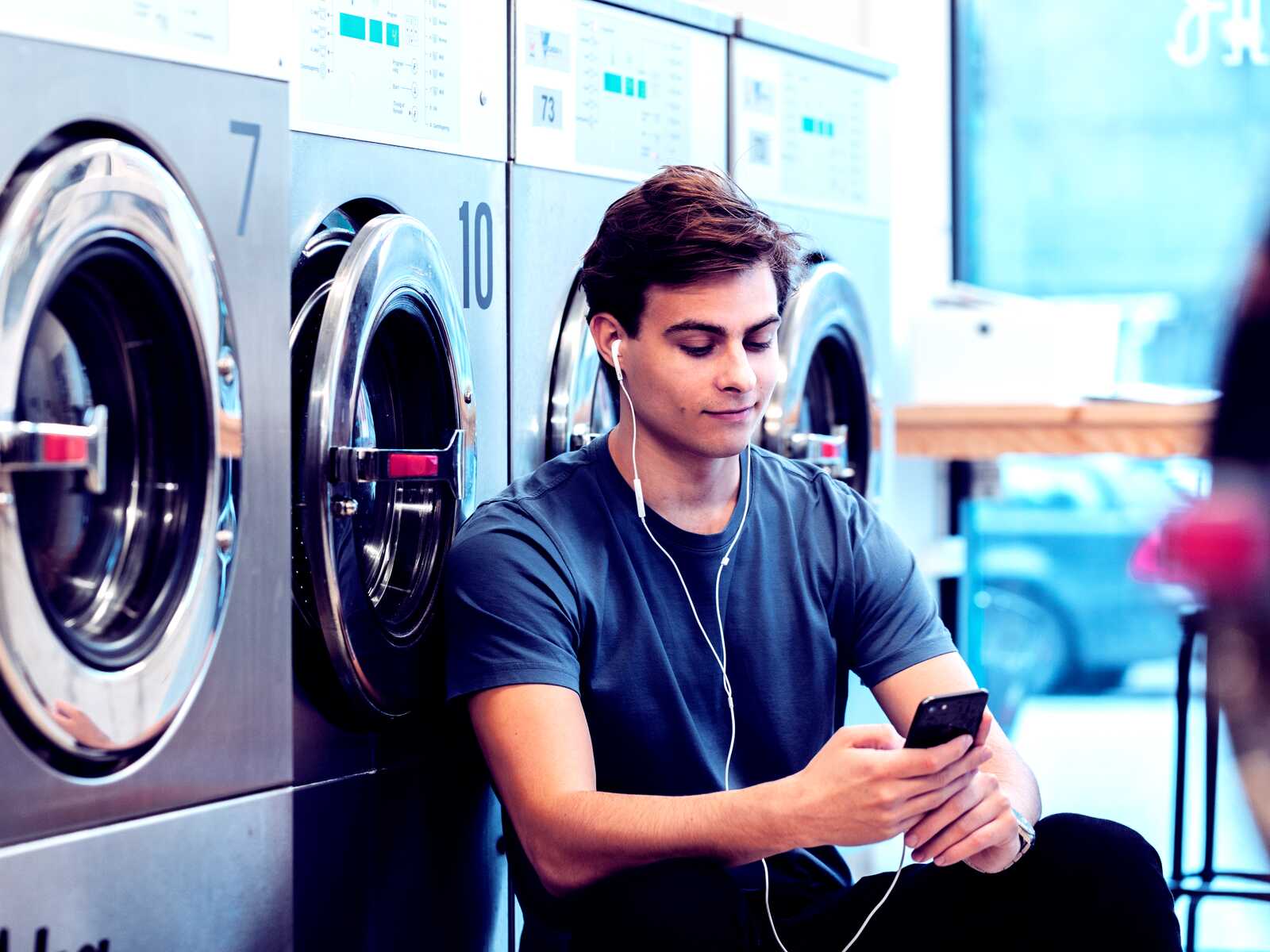 En ung mand sidder afslappet ved vaskemaskinerne og nyder musik, mens han tjekker sin telefon.