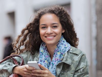 ung pige står udenfor med et stort smil og halstørklæde, men hun er ved at ordne noget på hendes telefon