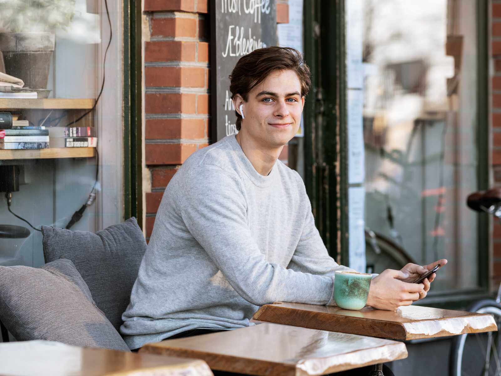 En ung mand sidder udenfor på en café og smiler til kameraet. Han har øretelefoner i og holder en telefon i hånden, mens der står en kop kaffe på bordet foran ham.