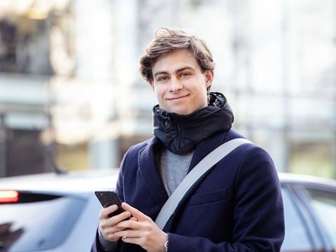 Ung mand iført mørk frakke smiler mod kameraet, mens han holder en telefon i hånden udendørs. Han har en afslappet og venlig fremtoning.