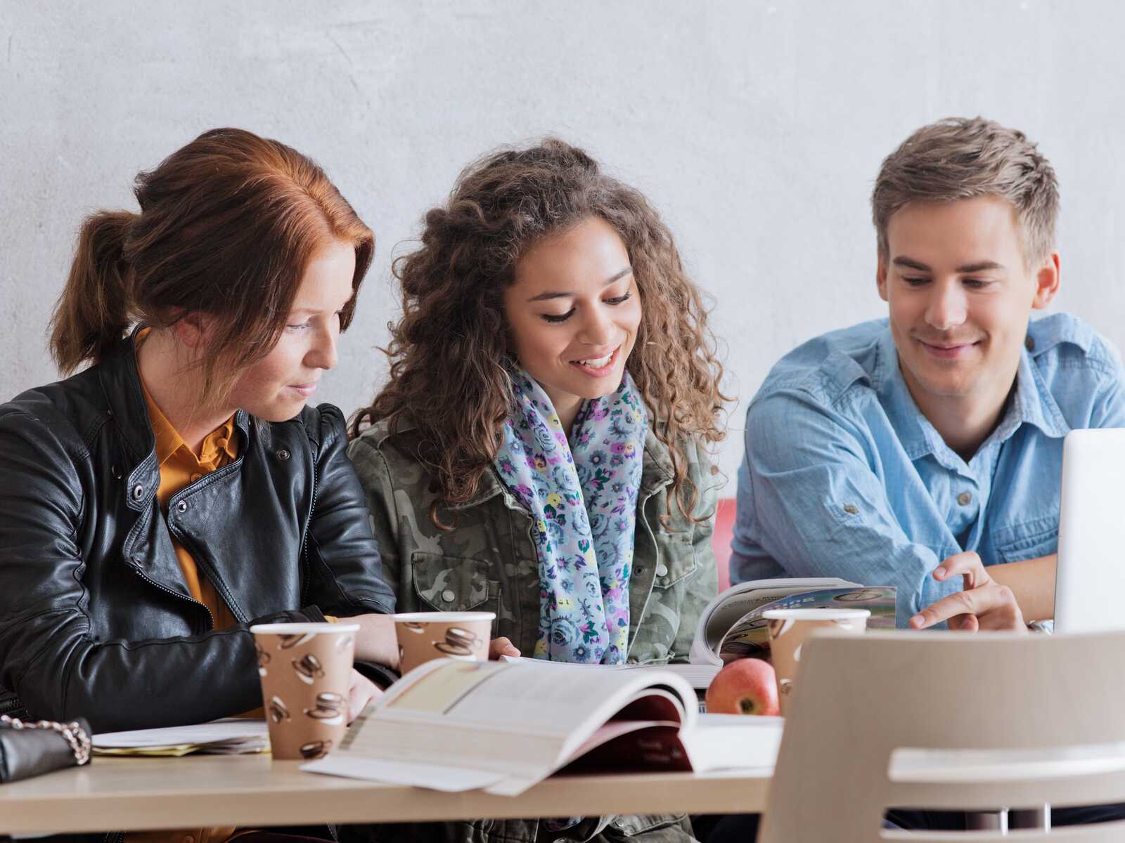 Tre unge studerende sidder sammen ved et bord og læser i bøger, mens de smiler og diskuterer. Kaffe og æbler står på bordet.