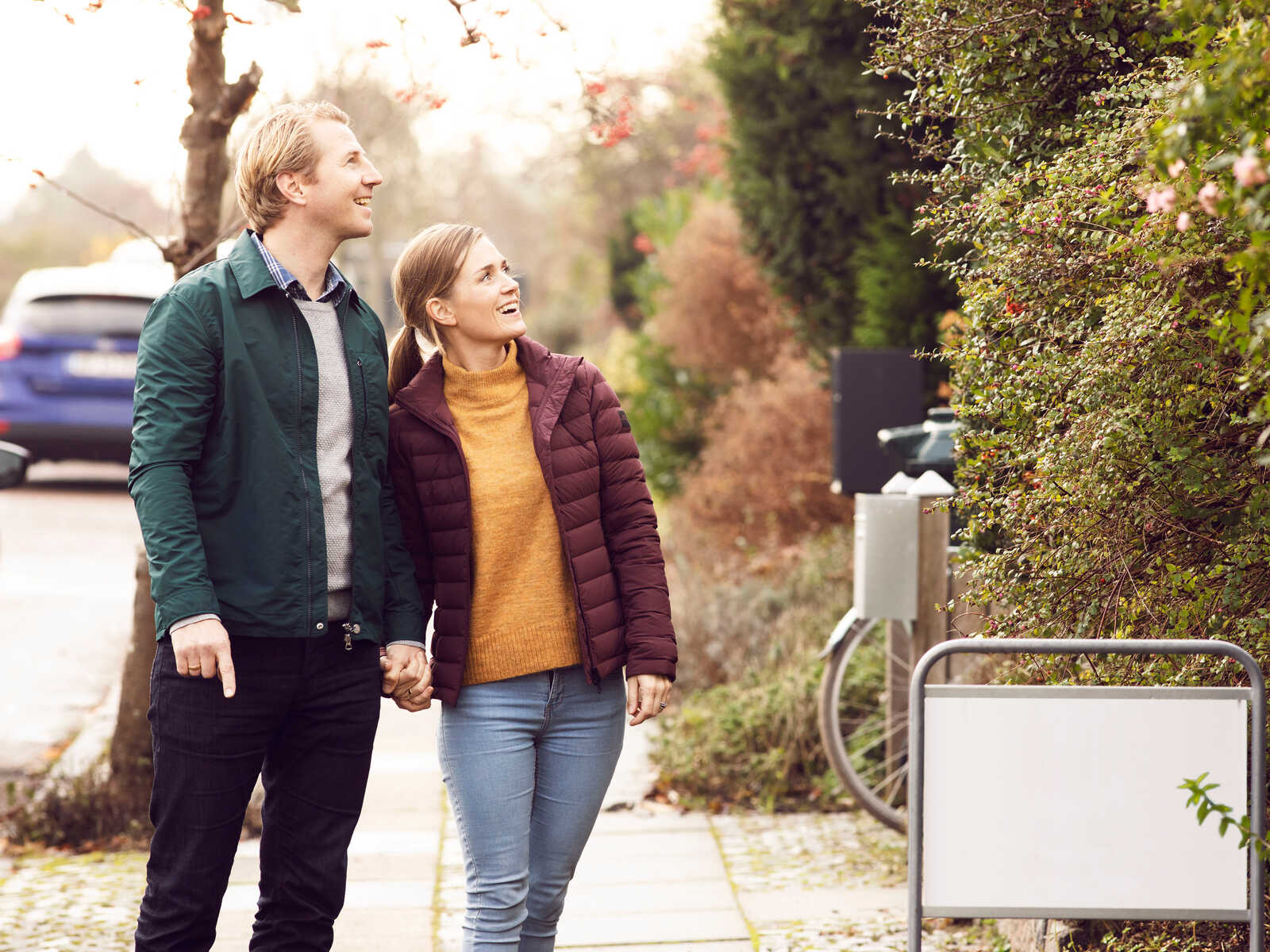 Ungt par går hånd i hånd i efterårsbeklædning, mens de smiler og kigger opad på hus.