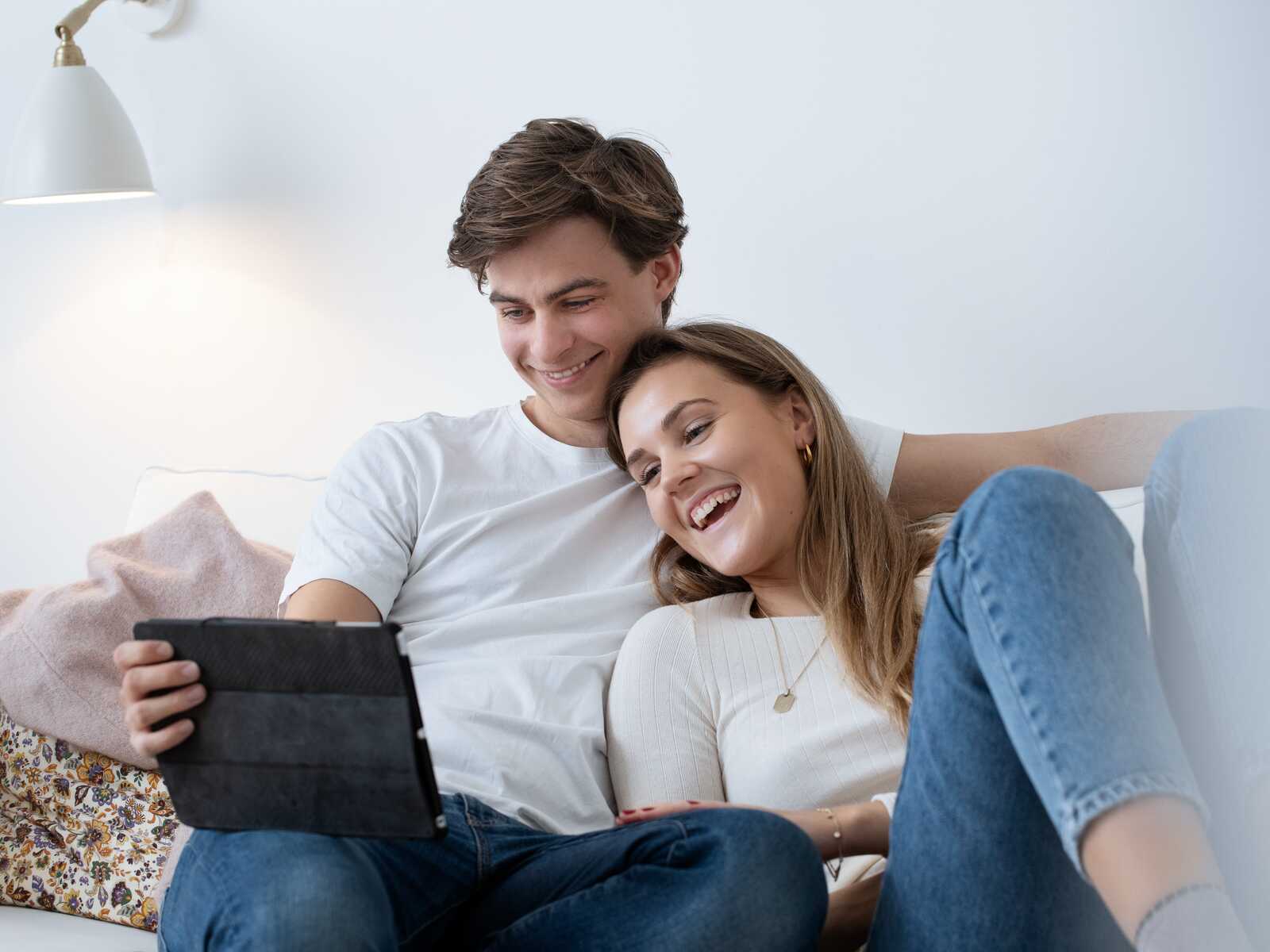 Ungt par sidder sammen i sofaen, griner og ser på en tablet, mens de er afslappede og smilende.