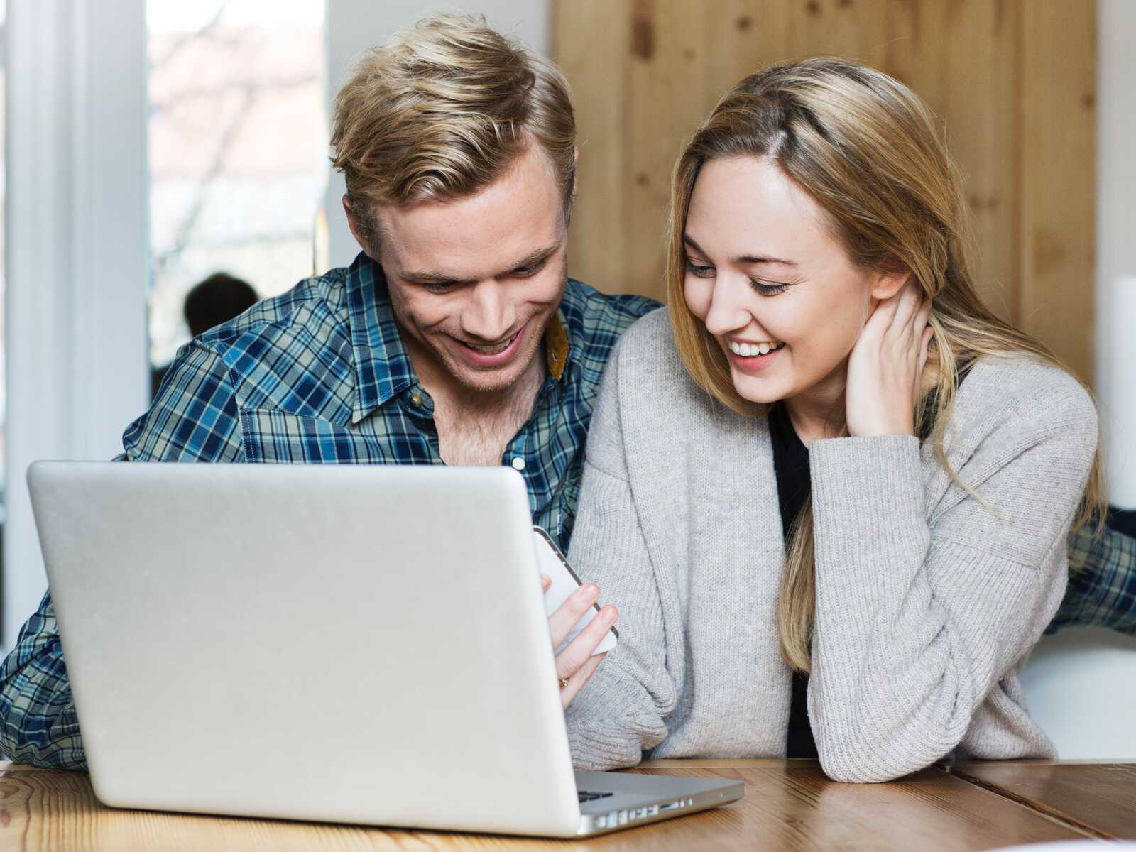 Et ungt par sidder ved et bord, ser på en laptop og en telefon og smiler.