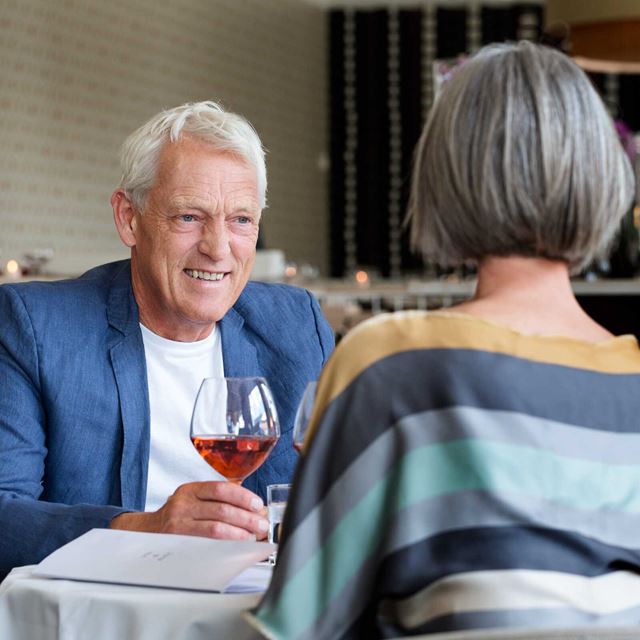 Manden smiler varmt med et glas vin i hånden, mens parret nyder en afslappet og intim middag i en elegant restaurant.