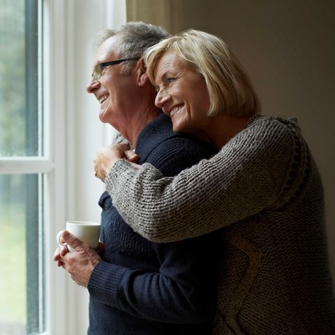 Ældre par står ved vinduet, kvinden krammer manden bagfra. Han holder en kop. Udendørs udsigt gennem vinduet, begge smiler varmt.