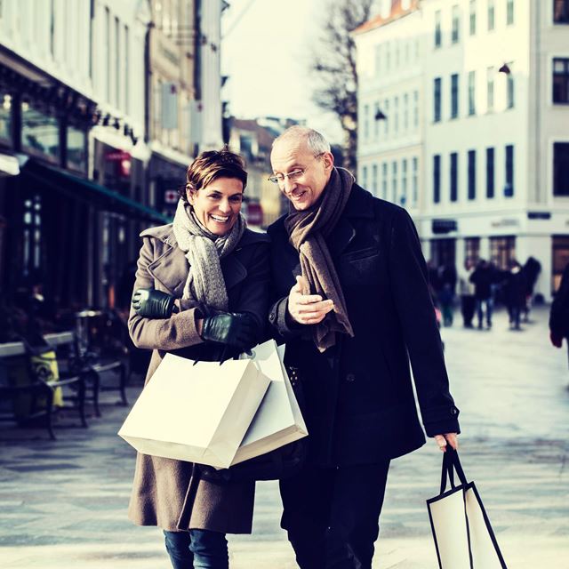 Ældre par går smilende ned ad en shoppinggade med indkøbsposer i hænderne. De er klædt i vintertøj, og gaden er fyldt med butikker.
