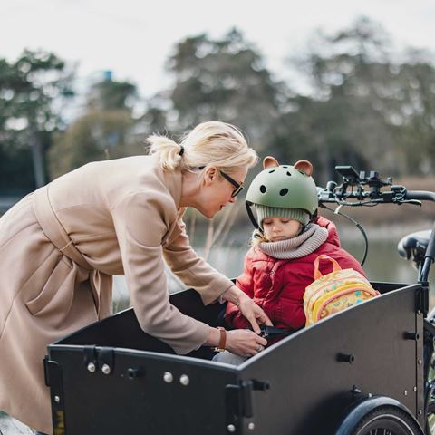 Mor hjælper sin datter med at spænde sikkerhedsselen i en ladcykel. Barnet bærer cykelhjelm og rygsæk, omgivelserne er udendørs med natur.