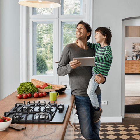 Far står i køkkenet med sin søn på armen og en tablet i hånden. Køkkenbordet er fyldt med friske grøntsager, og de kigger op på en lampe.