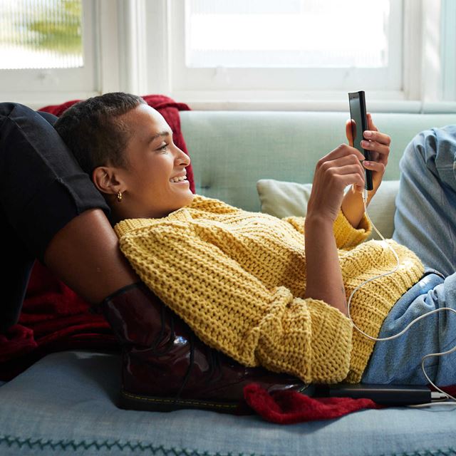 Kvinde i gul striktrøje smiler, mens hun ligger på sofaen og bruger en telefon med oplader i et hyggeligt opholdsrum.