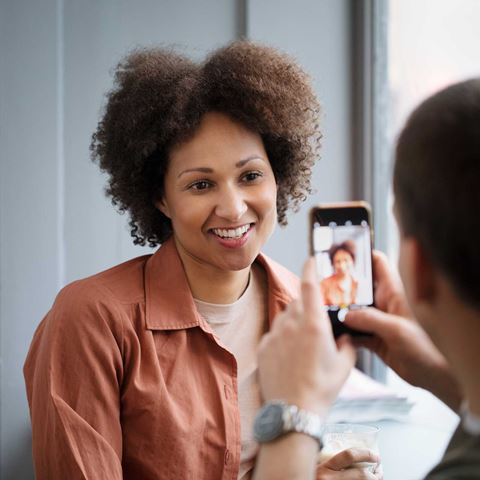 En kvinde smiler, mens hun får taget et billede med en telefon af en anden person i et lyst rum.