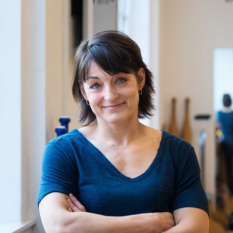 ældre dame i en blå t-shirt, står med armene over kors og smiler venligt ind i kameraret, baggrunden er sløret og beige