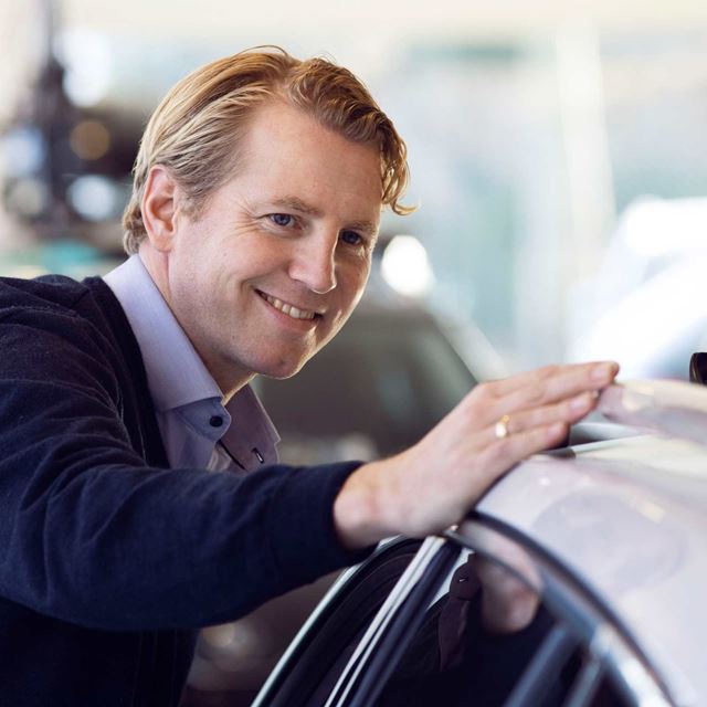 Smilende mand i forretningsdragt læner sig kærligt mod en ny bil i et showroom.