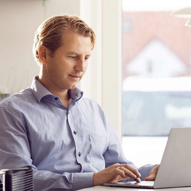 Mand i blå skjorte arbejder fokuseret på sin bærbare computer ved et vindue med naturligt lysindfald i baggrunden.