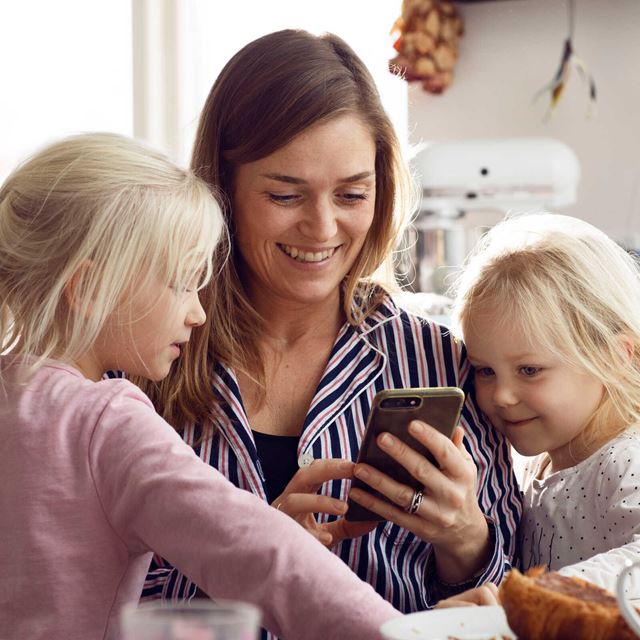 En mor sidder i køkkenet med sine to døtre, mens de alle smilende kigger på en smartphone sammen.