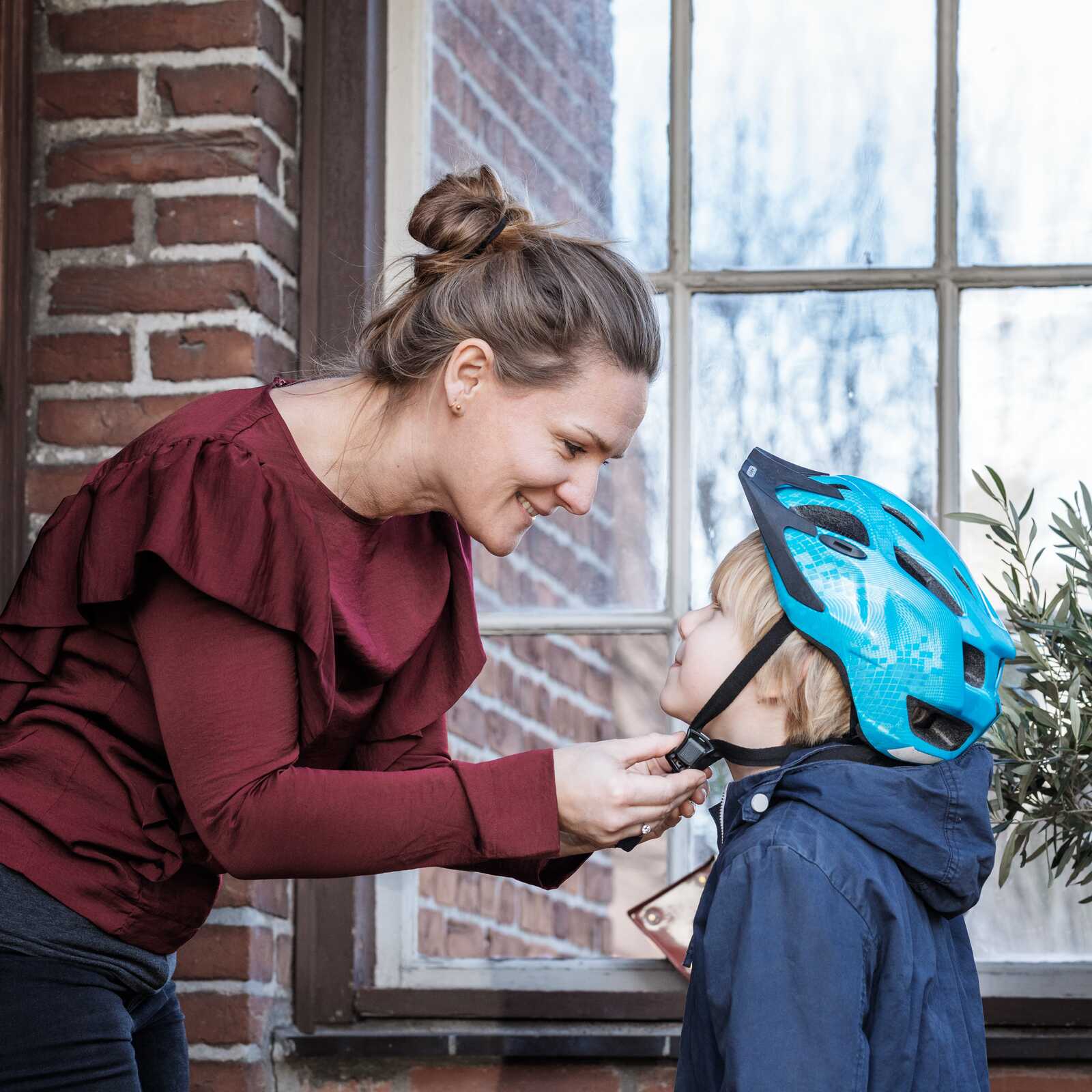 En mor fastgør sin søns cykelhjelm, mens de står udendørs foran et murstenshus og smiler til hinanden.