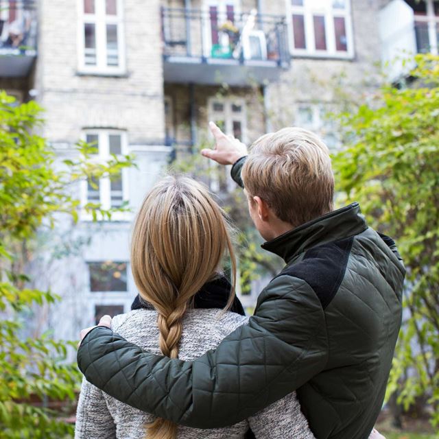 Et par står i en gårdhave og peger mod en lejlighedsbalkon, mens de kigger op på bygningen sammen.
